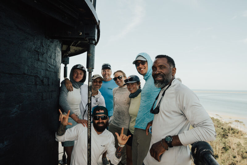 A group of eight people are posing for a photo on what appears to be a lighthouse balcony. They are dressed in casual clothing, with some wearing hats and sunglasses. The background features a scenic view of the ocean and trees. The overall atmosphere seems cheerful and relaxed.